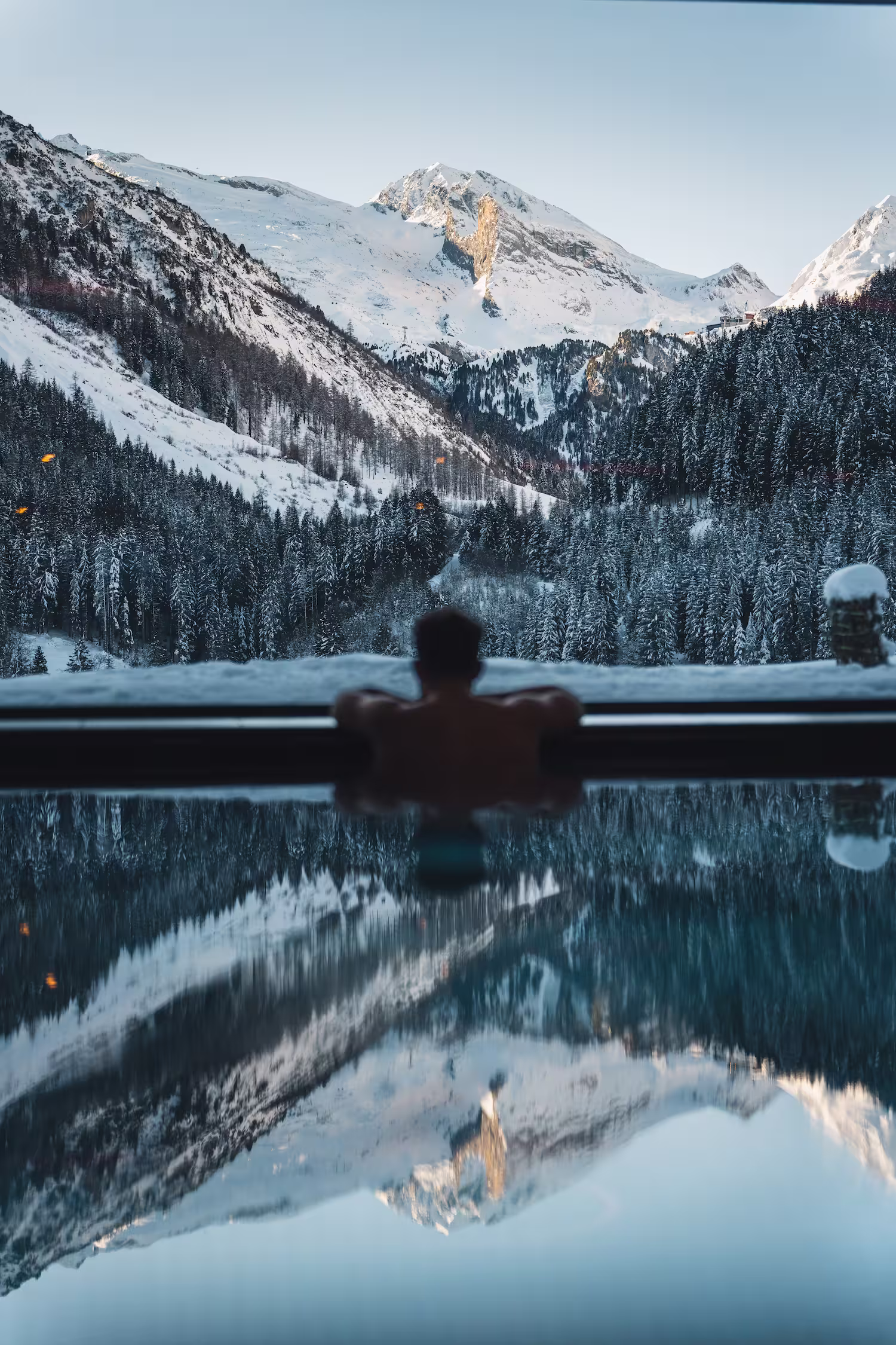 Person entspannt im Pool mit Blick auf verschneite Berge und Tannenwald in den Alpen.