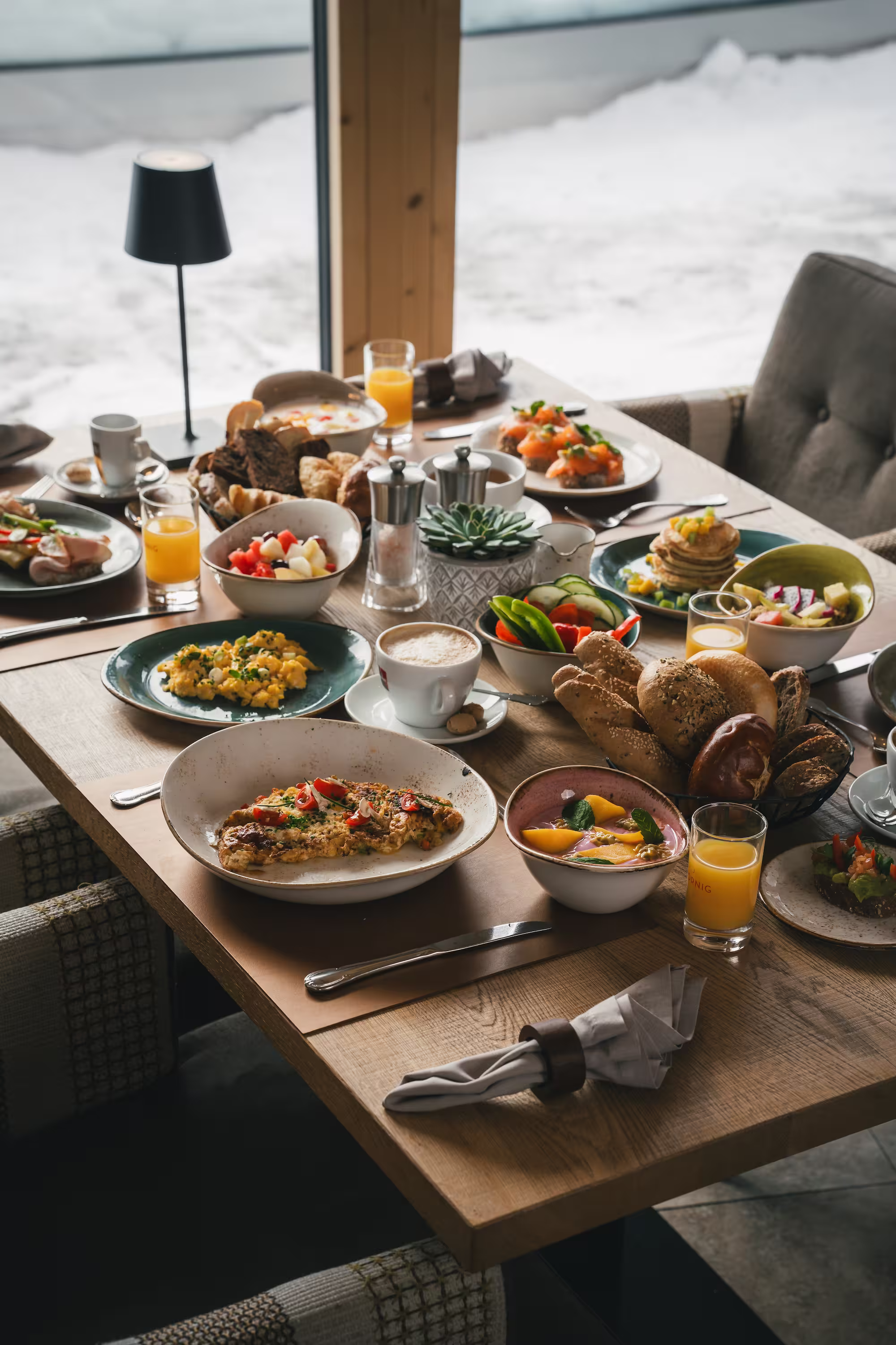 Gedeckter Frühstückstisch mit Rührei, frischem Obst, Gebäck und Orangensaft vor verschneiter Landschaft.