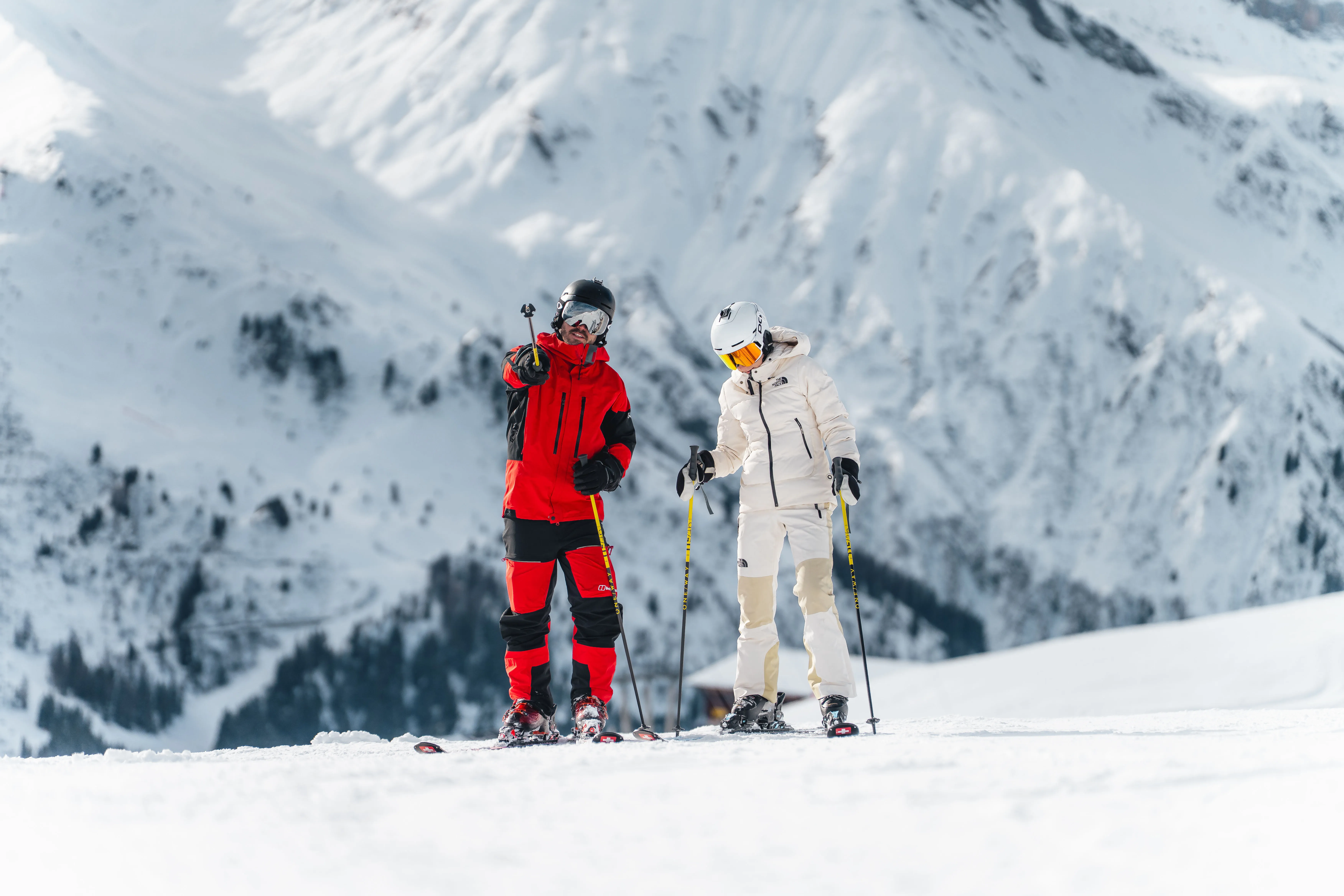 Zwei Skifahrer mit Helm und Skibrille vor einer verschneiten Bergflanke.