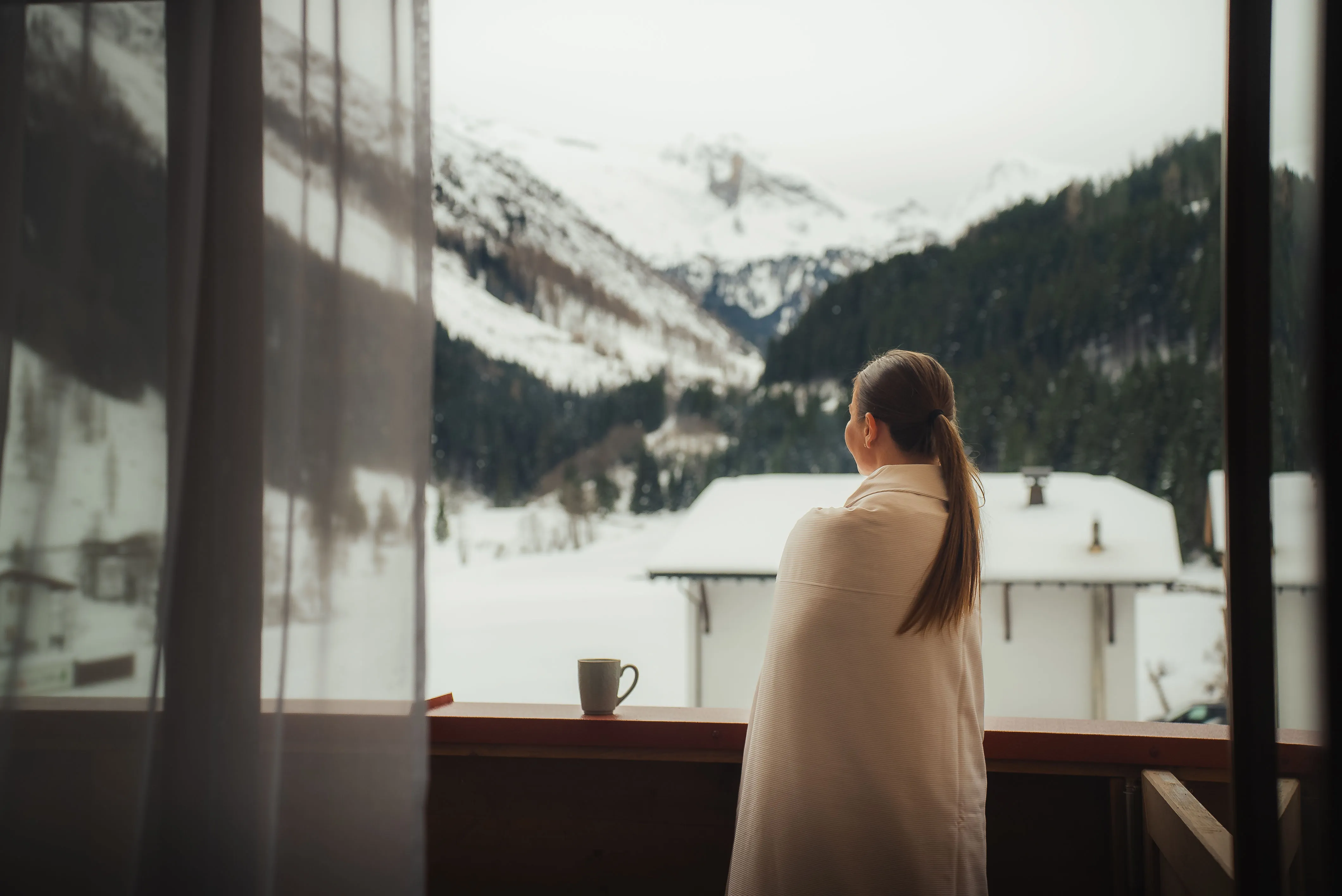Hotelgast in eine Decke gehüllt am Zimmerfenster mit Blick auf verschneite Berge.