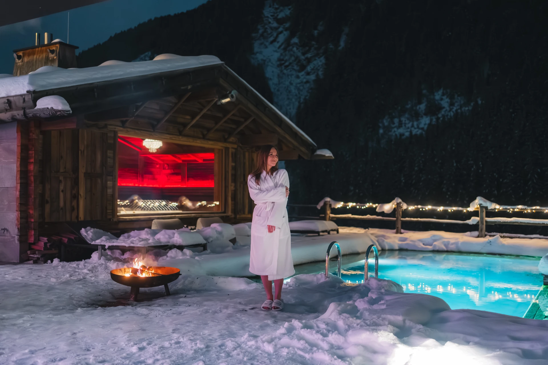 Hotelgast im Bademantel im Schnee vor dem beleuchteten Naturschwimmteich.