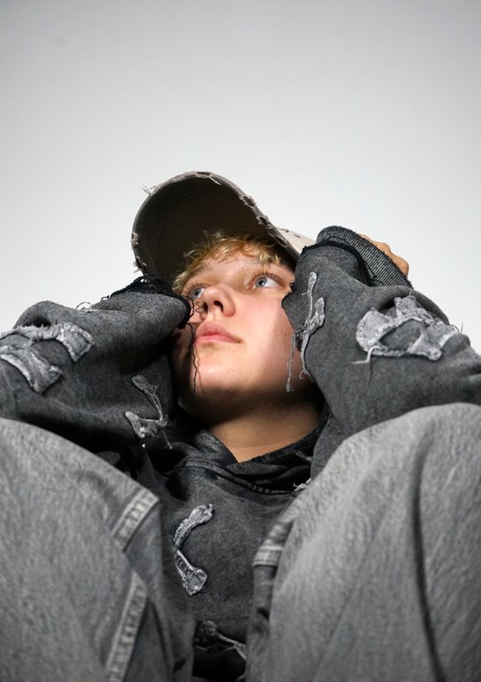 Young person with light curly hair wearing a distressed gray hoodie and matching pants, holding the brim of a light-colored cap while looking thoughtful against a plain light background.