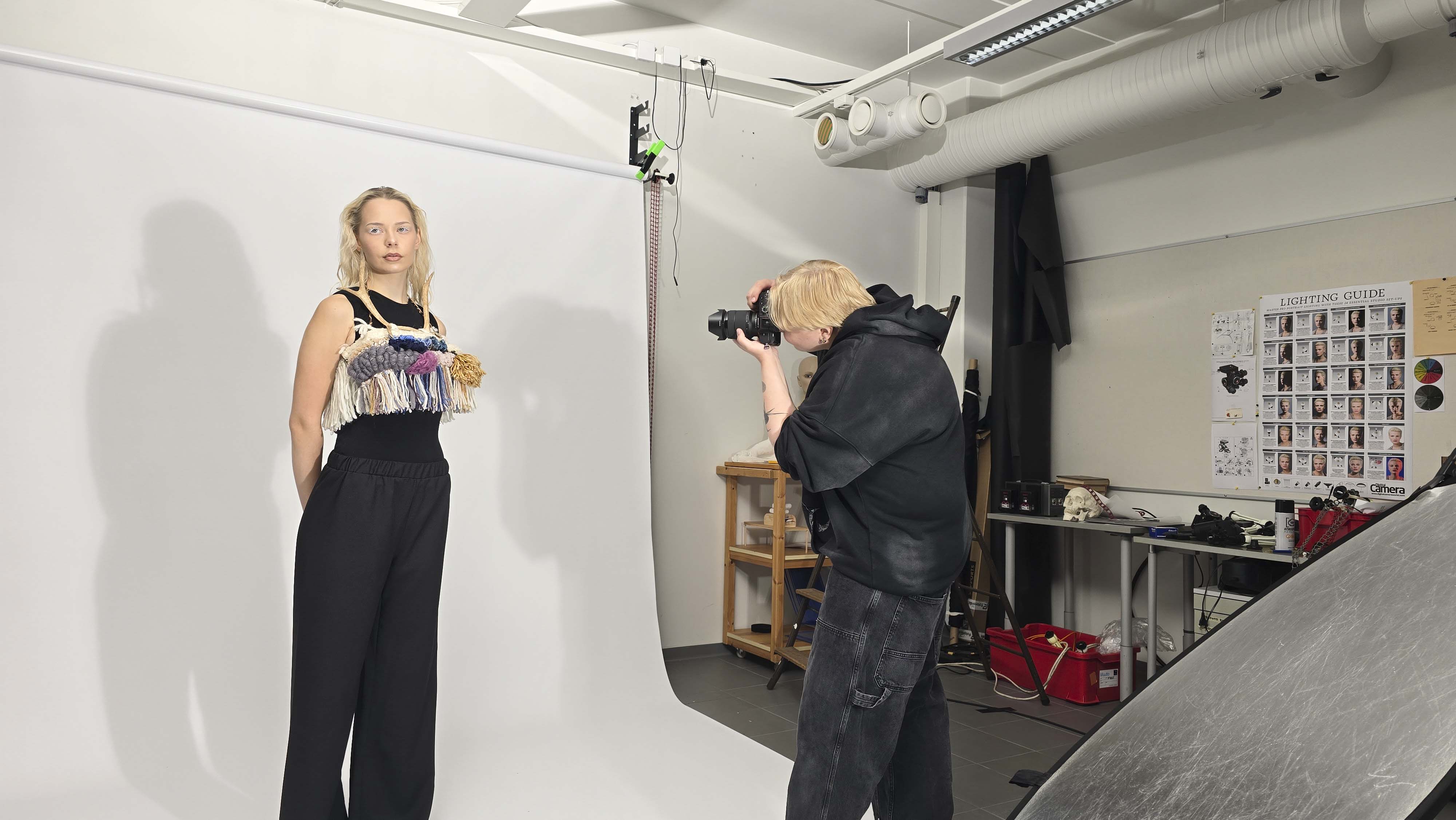 A photographer with short blond hair in black clothing takes photos of a female model in a studio with a white backdrop. The model wears black wide-leg pants and a sleeveless black top with colorful yarn embellishments on the chest. Studio equipment and a lighting guide poster are visible in the background.