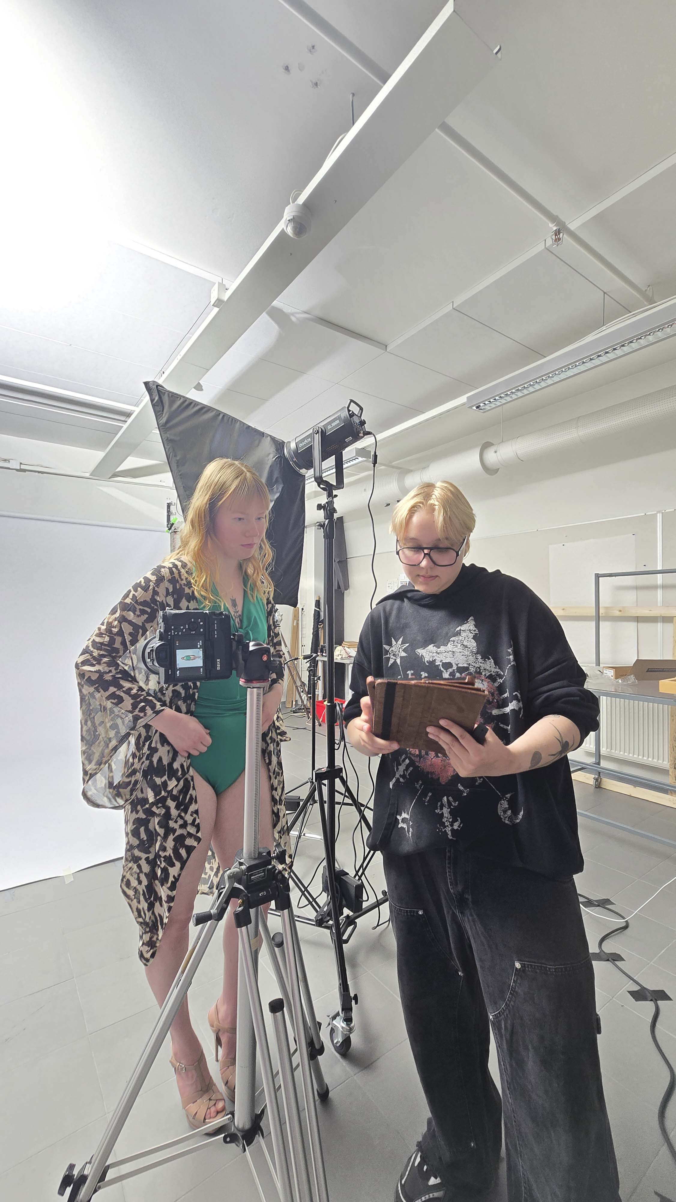 Two people in a photography studio; one person with long blonde hair wearing a green swimsuit and patterned cover-up stands behind a camera on a tripod, while the other with short blonde hair and glasses in a black hoodie and loose pants looks at a brown tablet device, surrounded by studio lights and equipment.