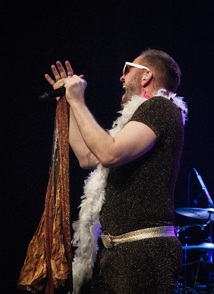 Male singer wearing a sparkly black outfit with a gold belt, white feather boa, and white sunglasses, passionately performing into a microphone on stage with a black background and drums in the back.