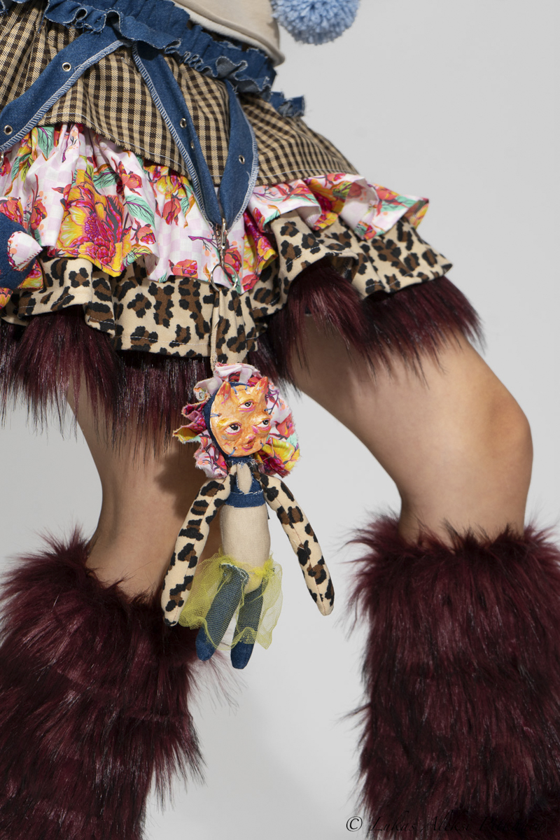 Person wearing a multi-patterned frilly skirt with layers of floral, leopard print, and checkered fabrics, attached with a small doll keychain featuring a three-eyed sun face and leopard print arms, paired with burgundy furry leg warmers.