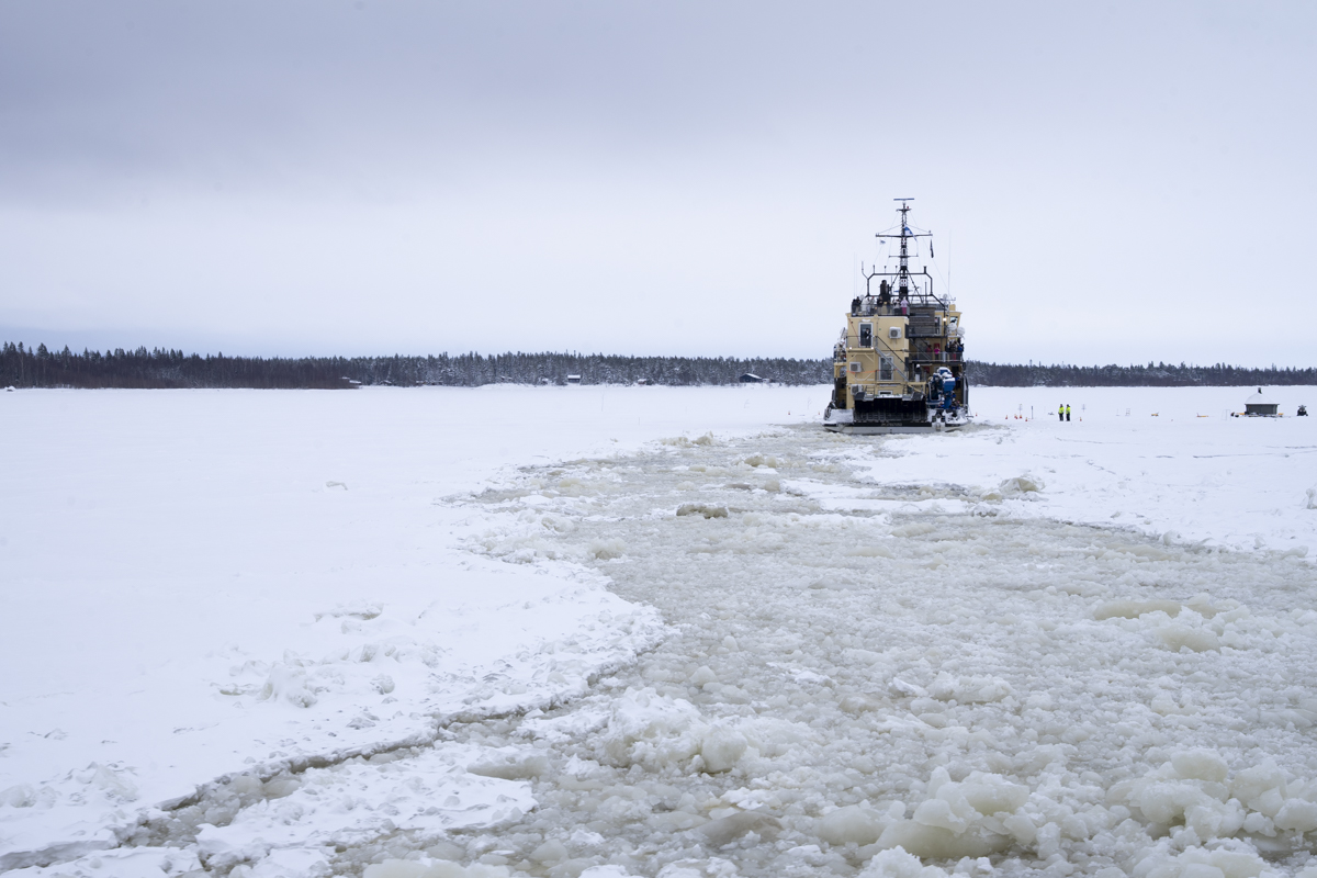 Icebreaker ship navigating through a frozen, snow-covered body of water, breaking a path in the thick ice under an overcast sky.