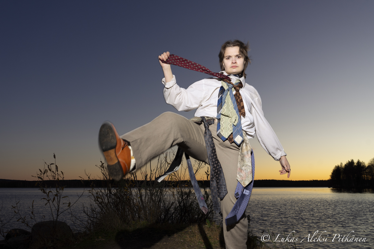Person standing outdoors near a lake at sunset, wearing multiple ties around the neck and waist, lifting one leg forward with a determined expression.