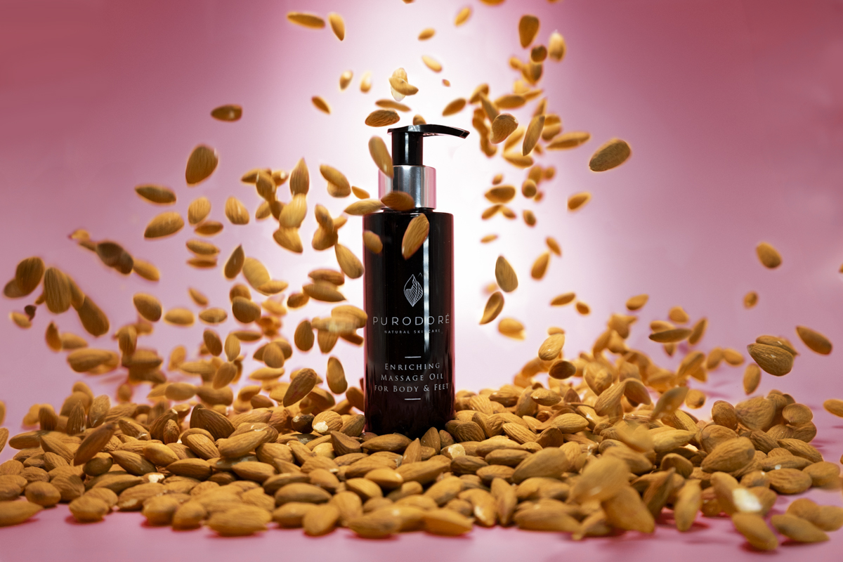 Dark bottle of Purodore enriching massage oil for body and feet surrounded by and mid-air almonds on a pink background.