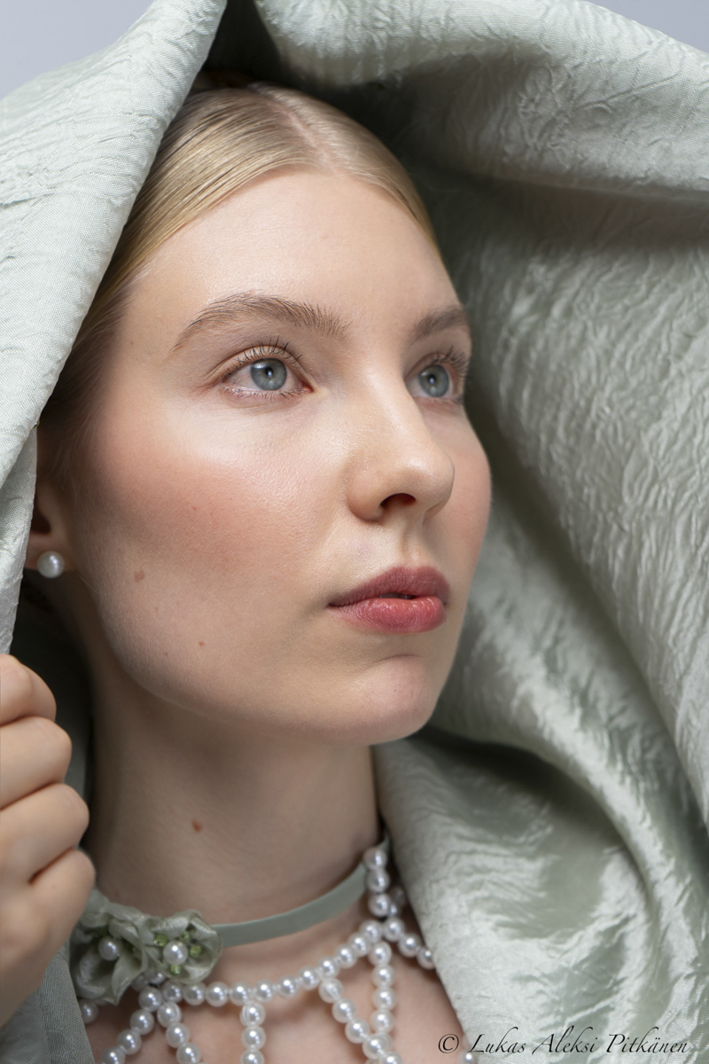 Close-up portrait of a young woman with light blue eyes and blonde hair, draped in a textured light green fabric, wearing pearl earrings and a layered pearl necklace.