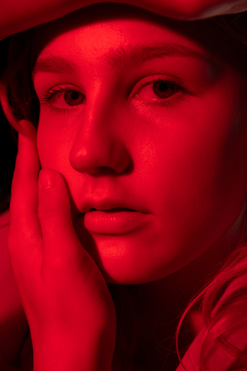 Close-up portrait of a young person with light skin and light-colored eyes, resting their face on their hand, illuminated by intense red lighting.
