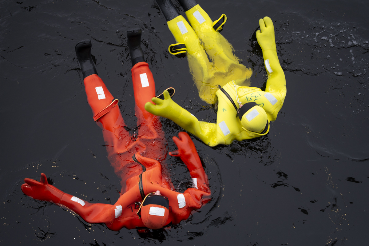 Two people floating on their backs in dark water wearing full-body survival suits, one in bright red and the other in bright yellow, with gloves and boots attached.