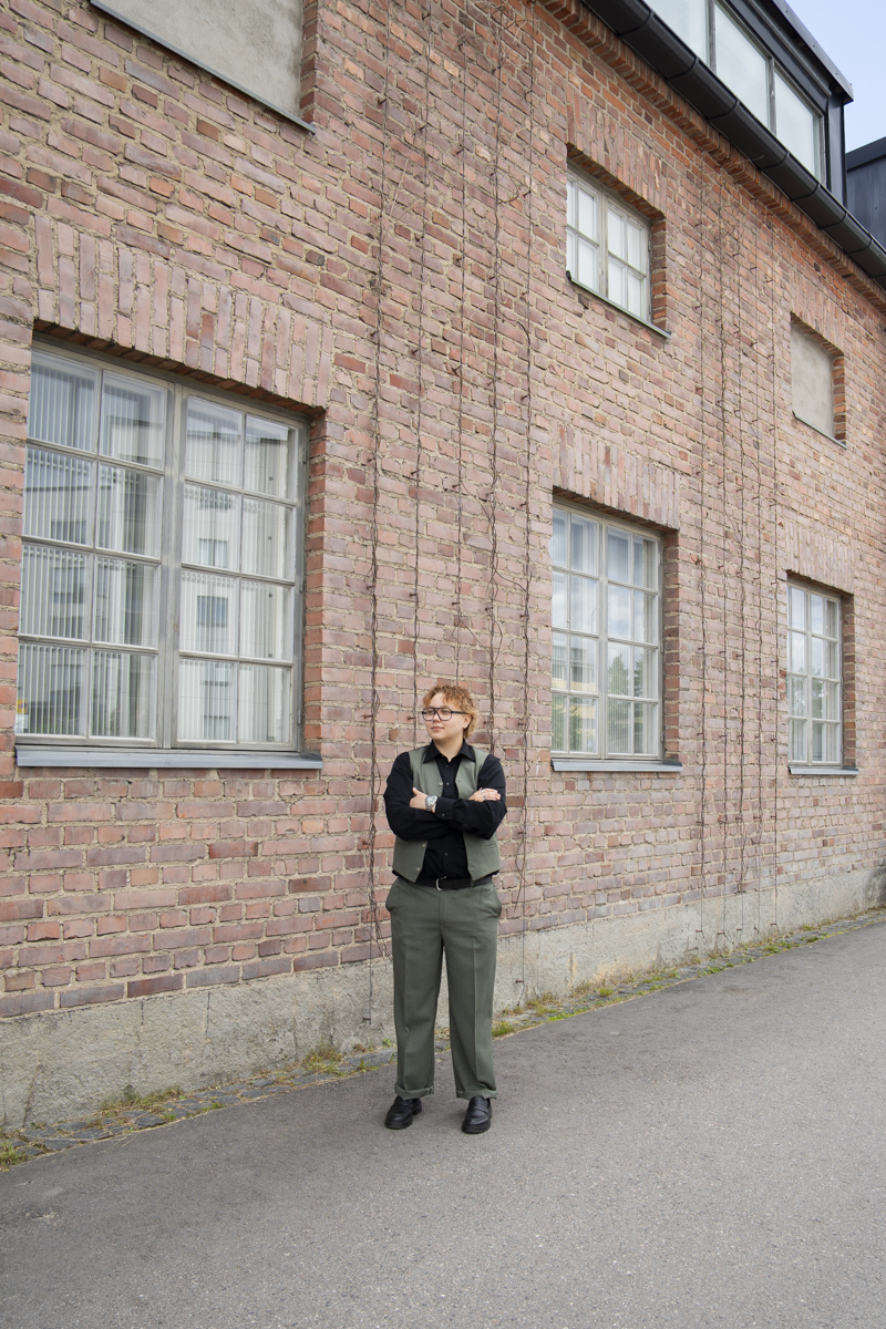 Person with short curly hair and glasses standing with arms crossed on a paved pathway against a brick building wall with large windows and climbing vines.