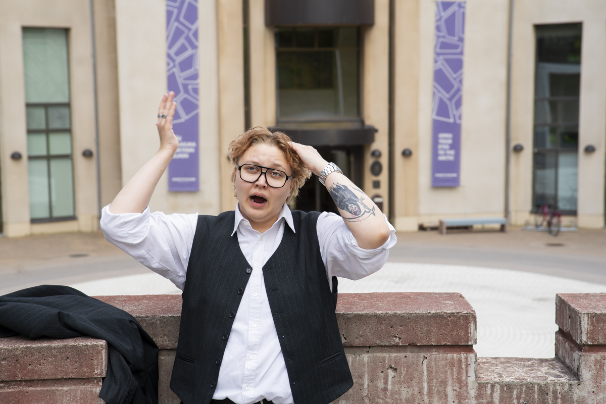Person with curly hair, wearing glasses and a black vest over a white shirt, stands outdoors in front of a building with light-colored walls and purple banners, raising one hand and touching their head with the other, with a tattoo visible on the left forearm.