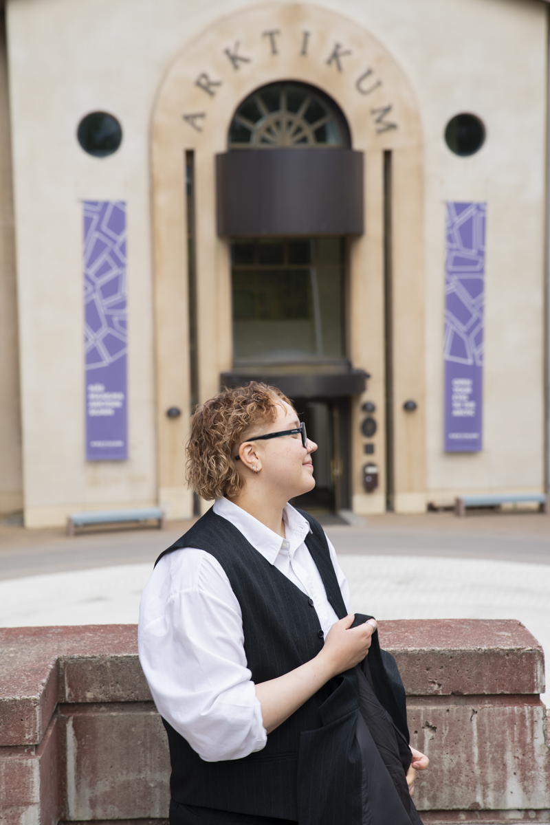 Person with short curly hair, glasses, and earrings wearing a white shirt and black vest standing near a brick ledge, looking to the right with a building entrance labeled 'ARKTIKUM' in the background.