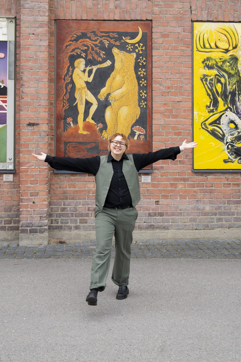 Person with curly blond hair, glasses, green vest and pants, and black shirt and shoes joyfully walking with arms outstretched in front of a brick wall with colorful framed artwork, including a central piece showing a person blowing a horn to a standing bear under a crescent moon.