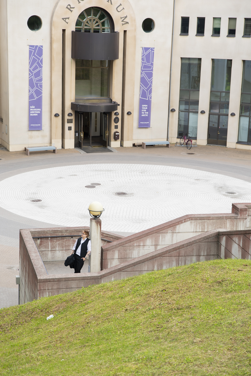 Person with short hair, wearing a white shirt and black vest, standing on stone steps near a round courtyard in front of a beige building entrance with two purple banners reading 'Open your eyes to the Arctic' and a bicycle parked nearby.