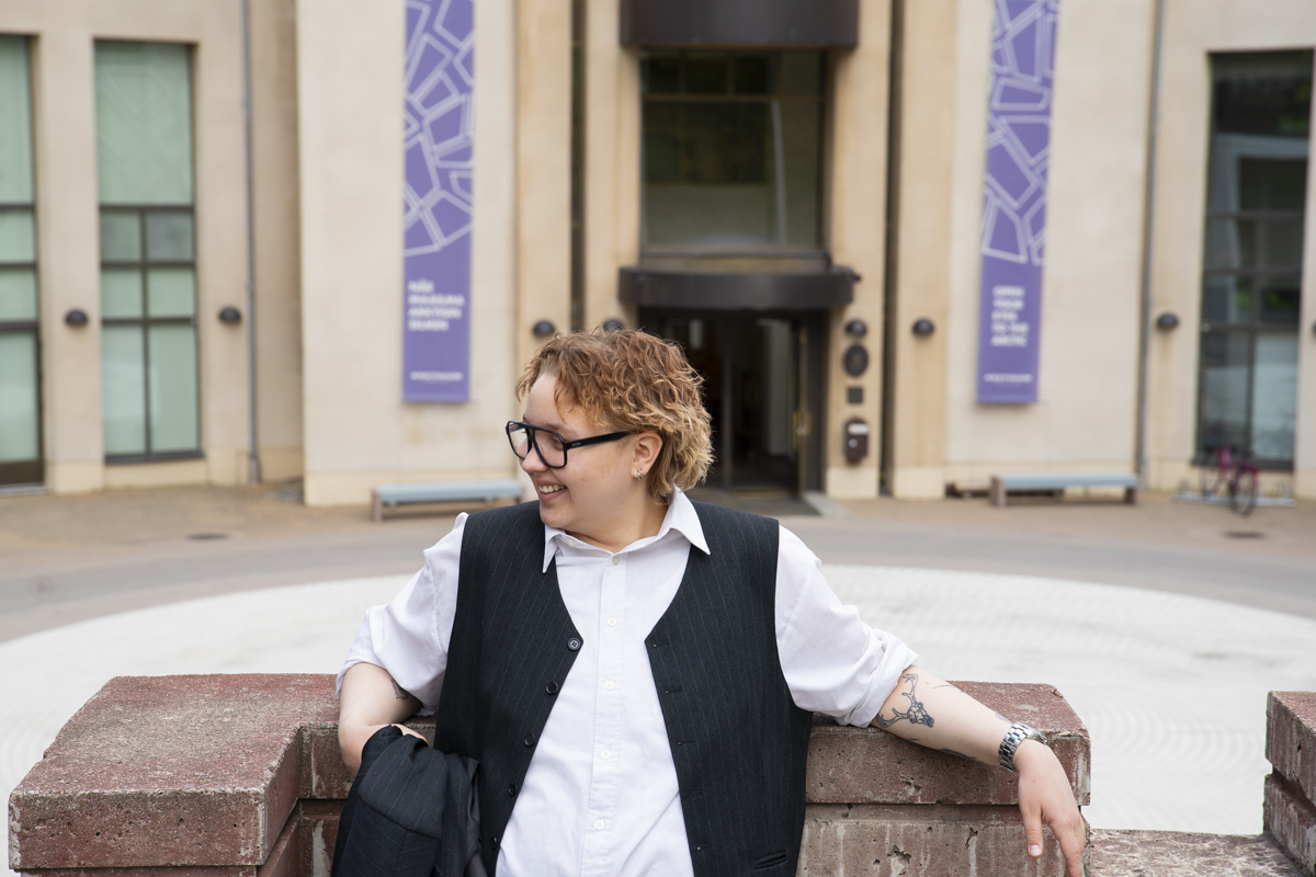 Person with short curly hair wearing glasses, a white shirt, and a dark vest leaning on a brick wall and smiling while looking to the side, with a building facade in the background.