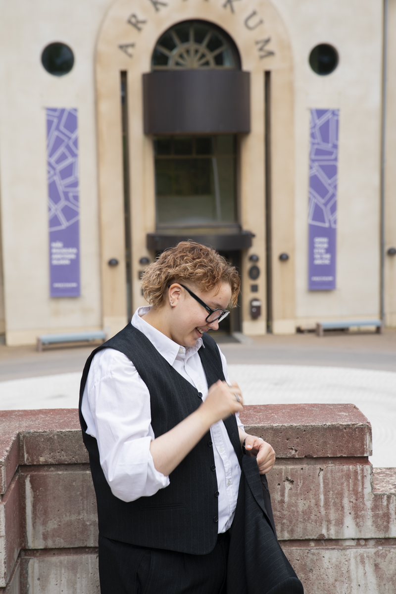 Person with curly short hair wearing black glasses, a white collared shirt, and a black vest standing outdoors near a stone wall with an arched building entrance in the background.
