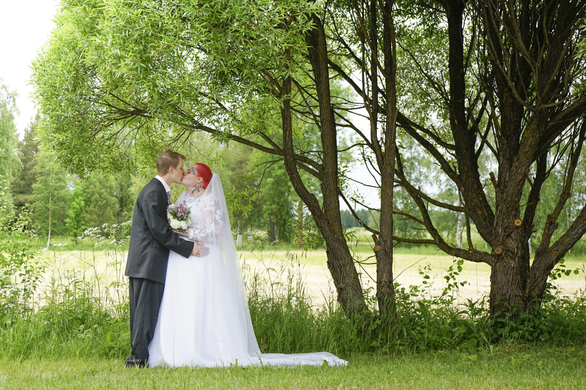 Bride and groom sharing a kiss outdoors under green trees, with the bride holding a bouquet and wearing a white dress and veil, and the groom in a dark suit.