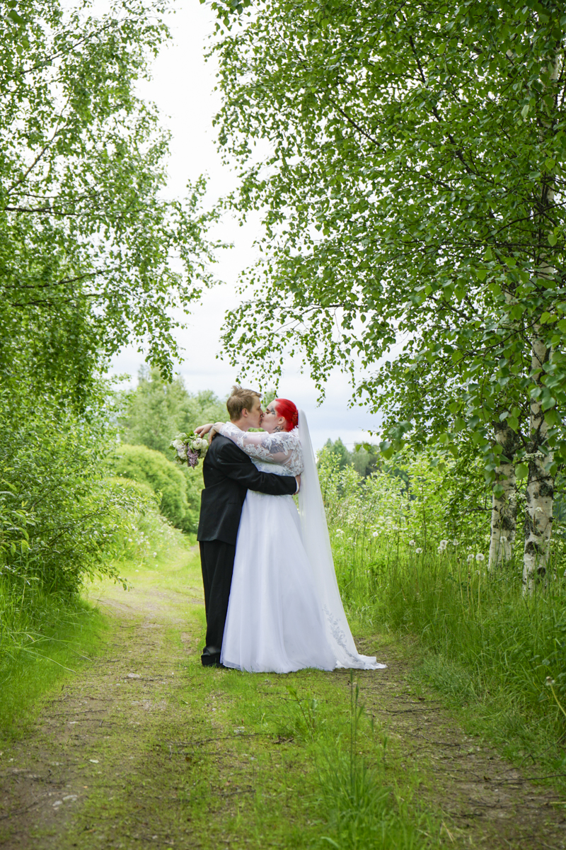 Bride with bright red hair in a white wedding gown and veil kissing groom in a black suit while holding a bouquet, standing on a green leafy path surrounded by tall trees.