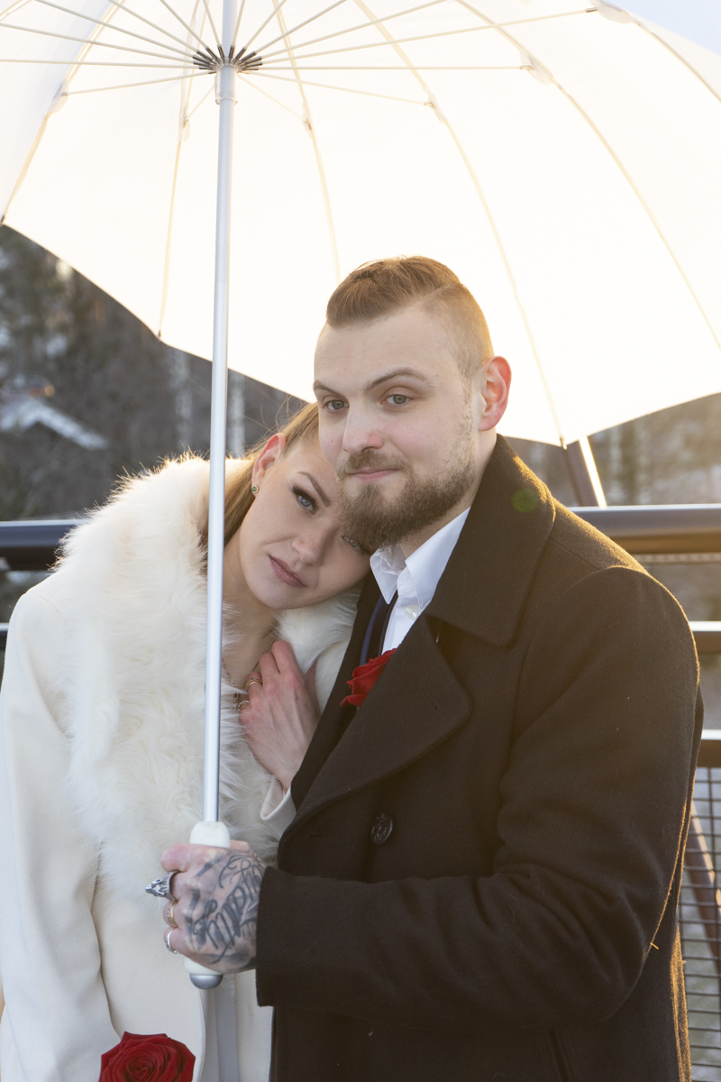 A couple standing close under a white umbrella, the woman in a white fur coat resting her head on the man's shoulder, the man wearing a black coat with a red rose on his chest, holding the umbrella with a tattooed hand, both looking content.