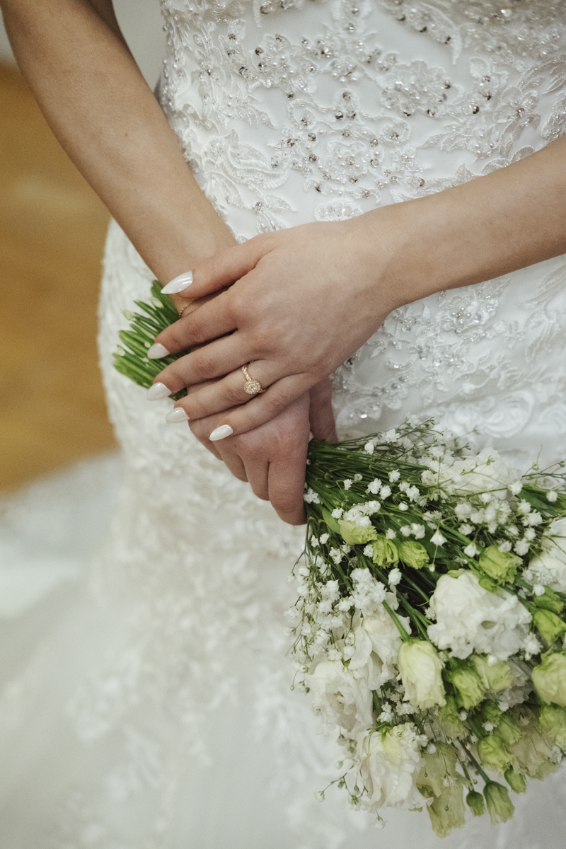 Close-up of a bride's hands with a gold engagement ring holding a bouquet of white and green flowers, wearing a white embroidered wedding dress.