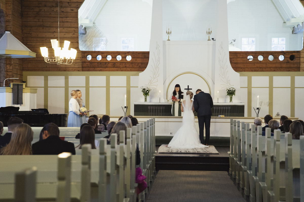Bride and groom standing at the altar in a church with a minister officiating the wedding ceremony, guests seated in pews facing them, bridesmaids holding bouquets to the left.