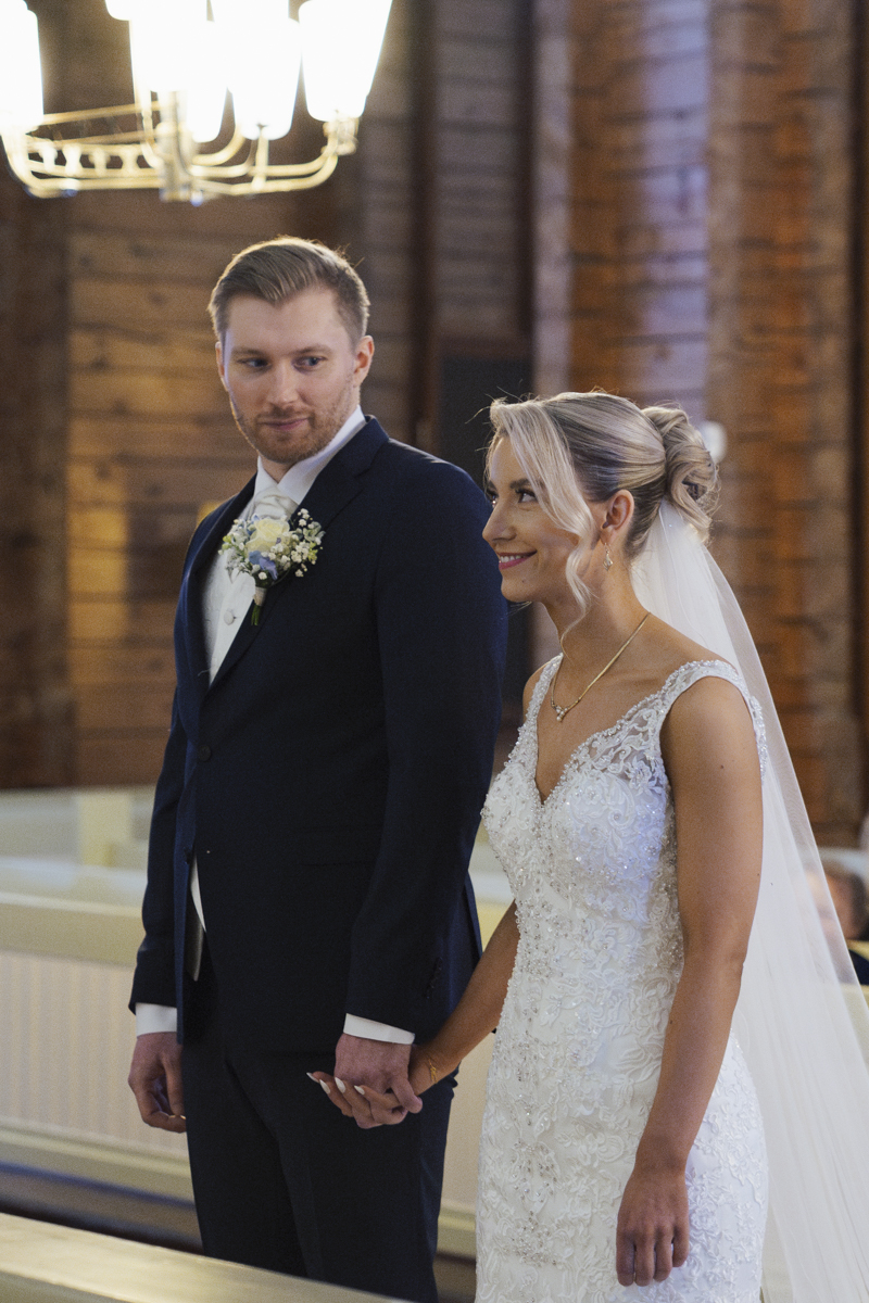 Bride in a white lace wedding dress and veil holding hands with groom in a dark suit with a boutonniere, standing indoors with wooden walls and a chandelier in the background.