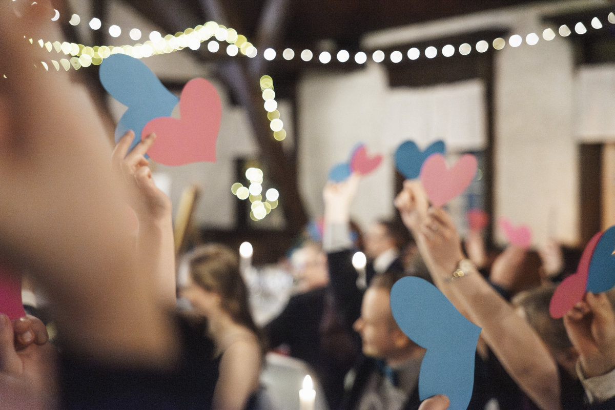 Guests at a formal event holding up pink and blue paper hearts, with blurred background and string lights overhead.