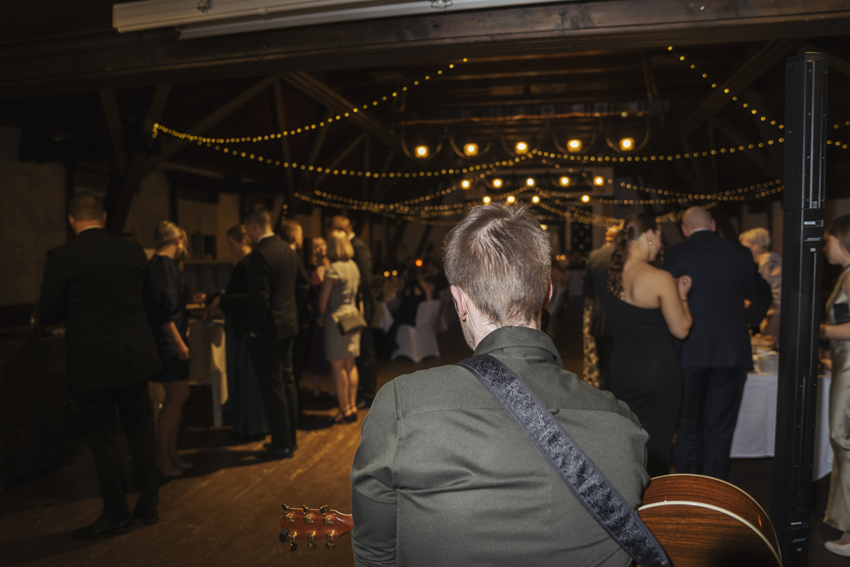 View from behind a musician playing guitar at a dimly lit indoor event with string lights overhead and guests dressed formally standing and mingling.