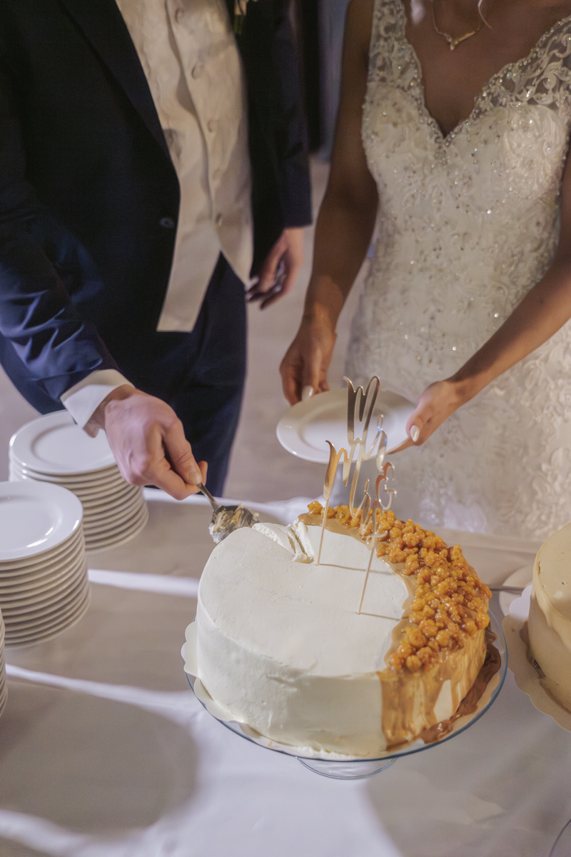 A bride and groom cutting a wedding cake with white frosting and topped with caramel popcorn and a golden Mr & Mrs cake topper, with stacks of white plates nearby.
