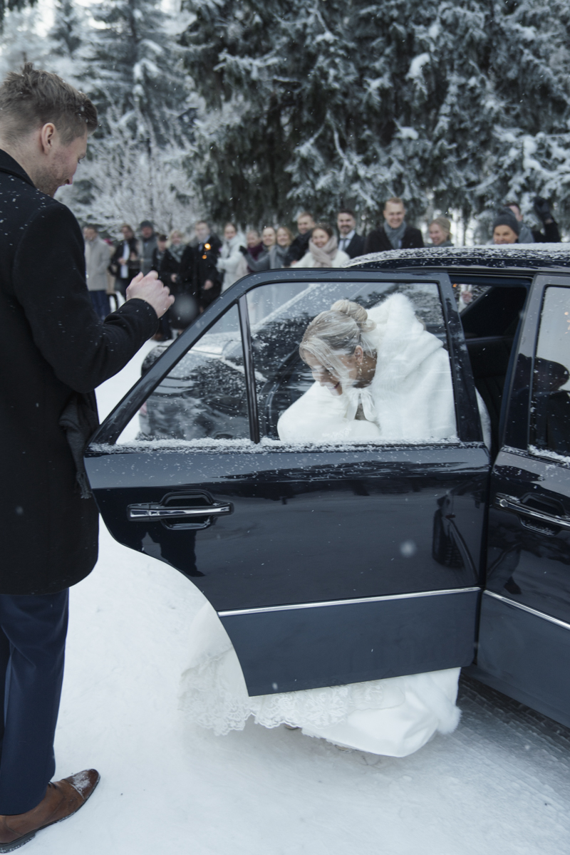 Bride wearing a white fur coat and wedding dress getting out of a black car in snowy outdoor setting while a man in a dark coat and brown shoes stands nearby, with smiling guests and snow-covered trees in the background.