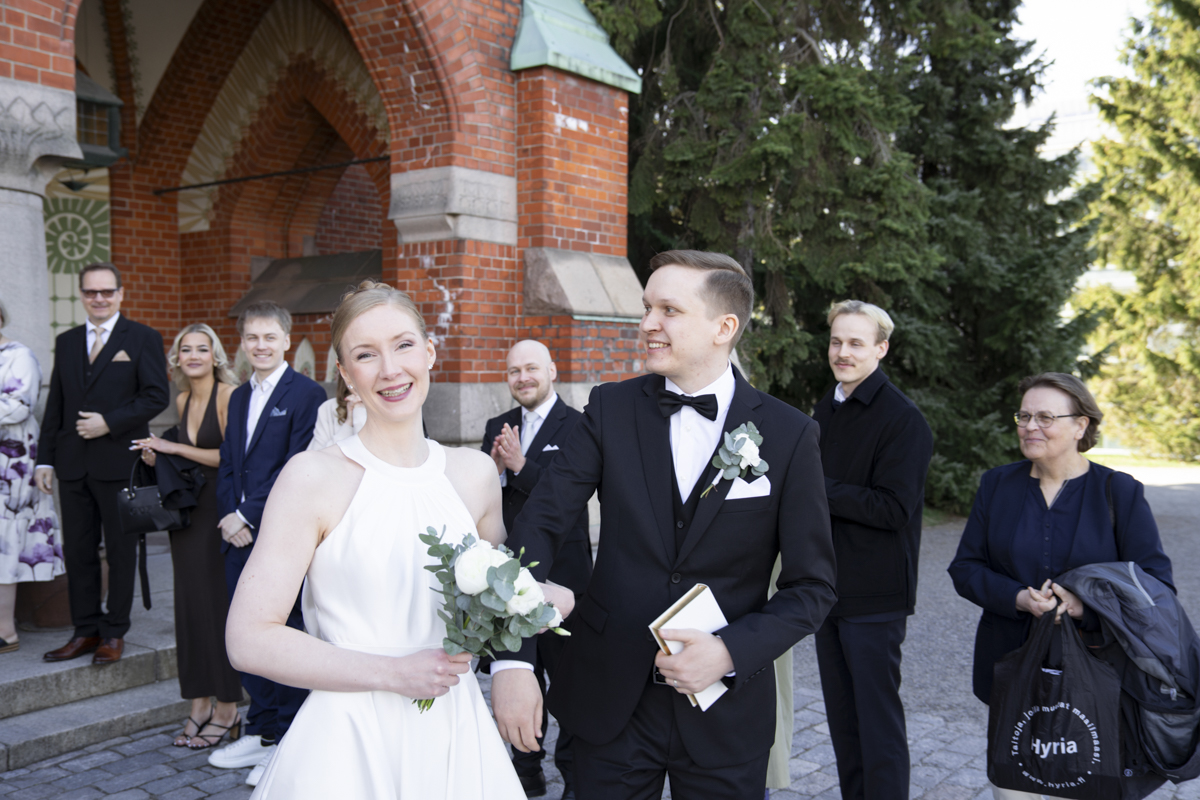 A bride in a white sleeveless dress holding a bouquet of white flowers and a groom in a black tuxedo with a bow tie and boutonnière smile and hold hands outside a brick building with guests standing behind them, some clapping and smiling.