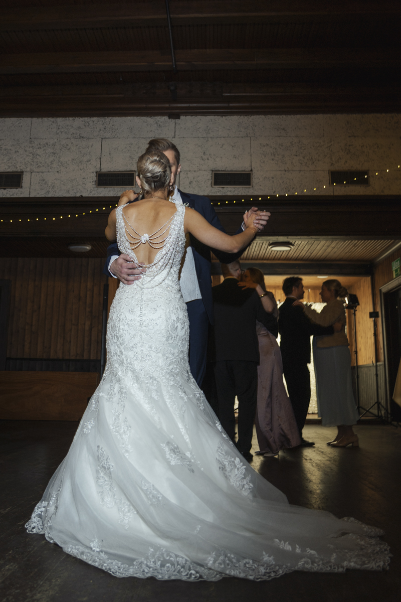 Bride in a white lace wedding gown with an open back decorated with pearl chains dancing with the groom in a dark suit in a rustic wood-paneled room with other couples dancing in the background.