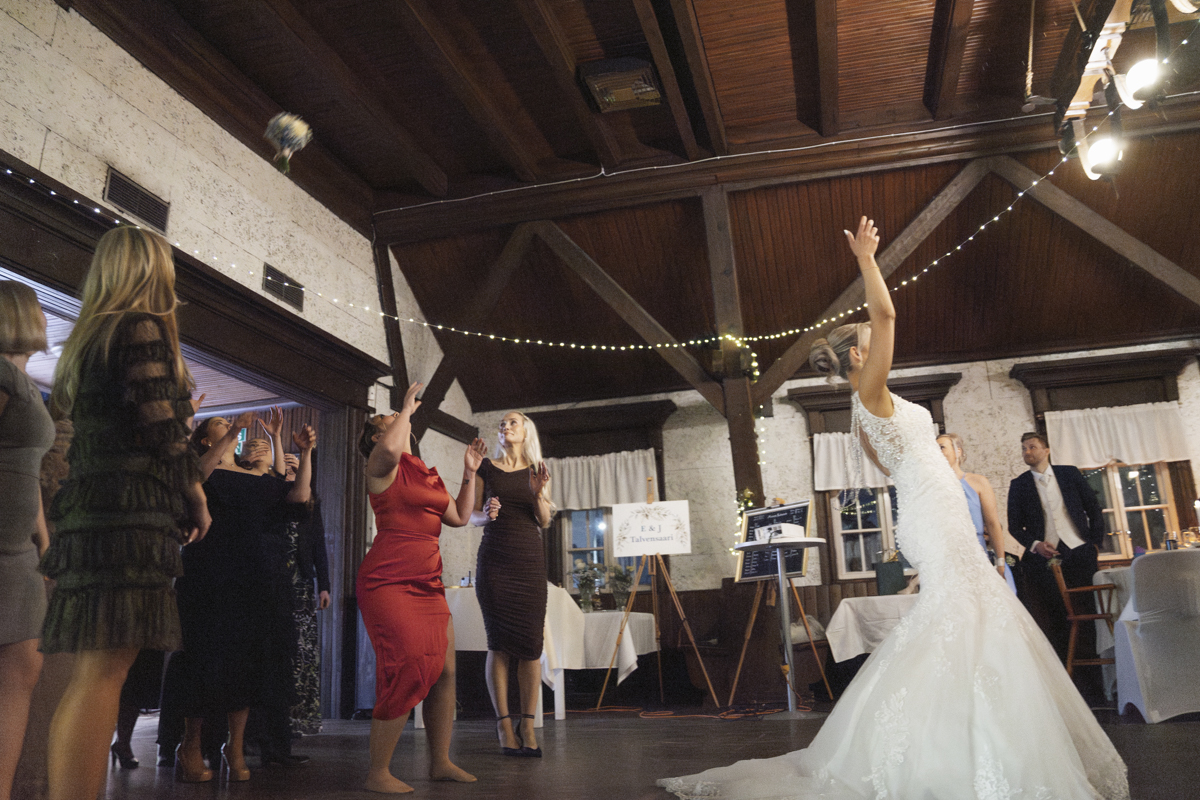 Bride in white lace wedding dress tossing bouquet over her shoulder towards group of women eagerly reaching up to catch it in a rustic venue with wooden beams and string lights.