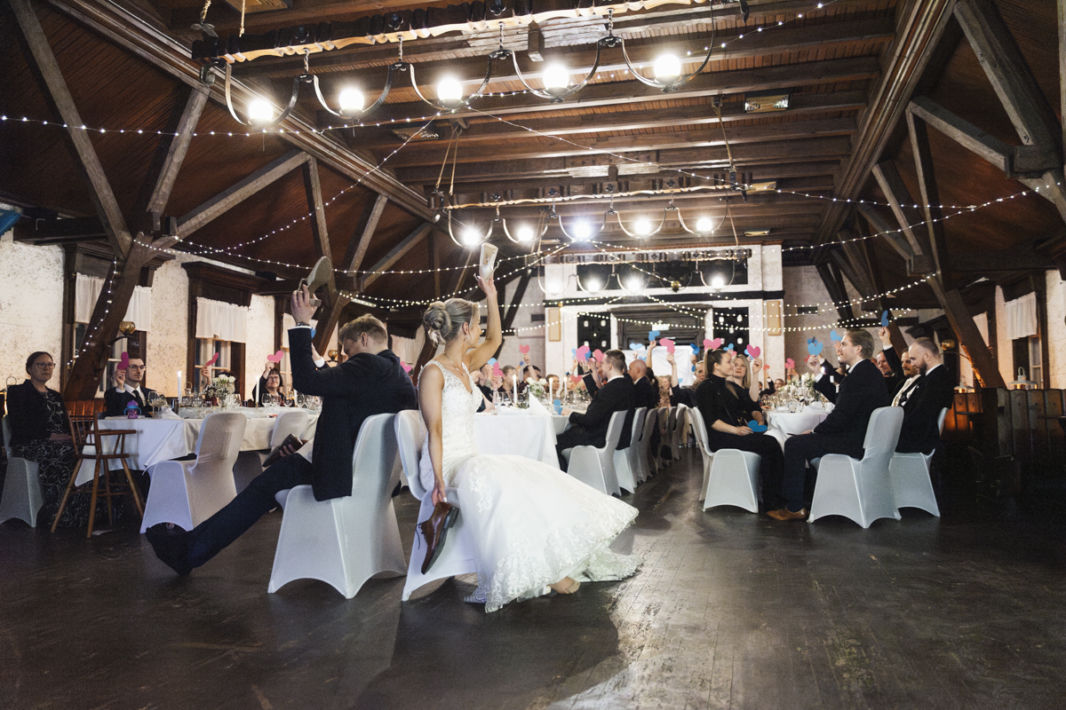 Indoor wedding reception with guests seated at decorated tables raising pink and blue heart-shaped cards, a bride in a white gown and groom in a suit sitting back to back holding each other's shoes.