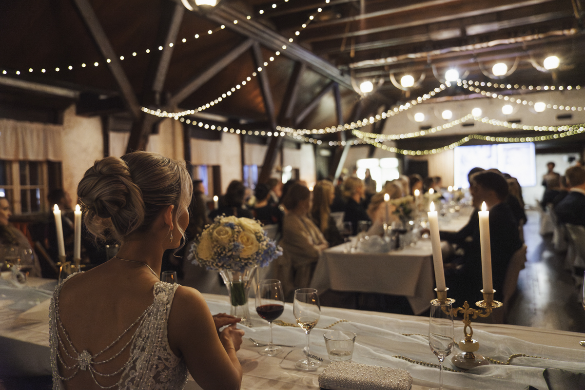 Bride with an elegant updo hairstyle and beaded wedding dress sitting at a decorated table with candles and floral centerpiece, facing guests seated at long banquet tables in a wooden venue with string lights overhead.