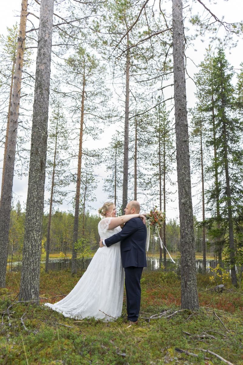 Bride in a white lace wedding dress and groom in a dark suit embracing in a forest clearing with tall pine trees and a small pond in the background.