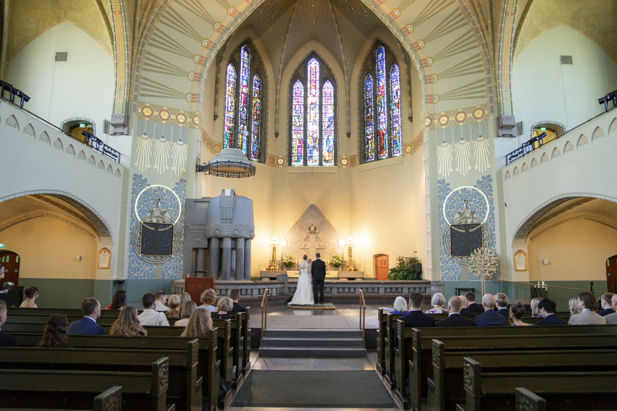 A wedding ceremony inside a church with a bride and groom standing at the altar, guests seated in wooden pews, and tall stained glass windows illuminating the decorated interior.