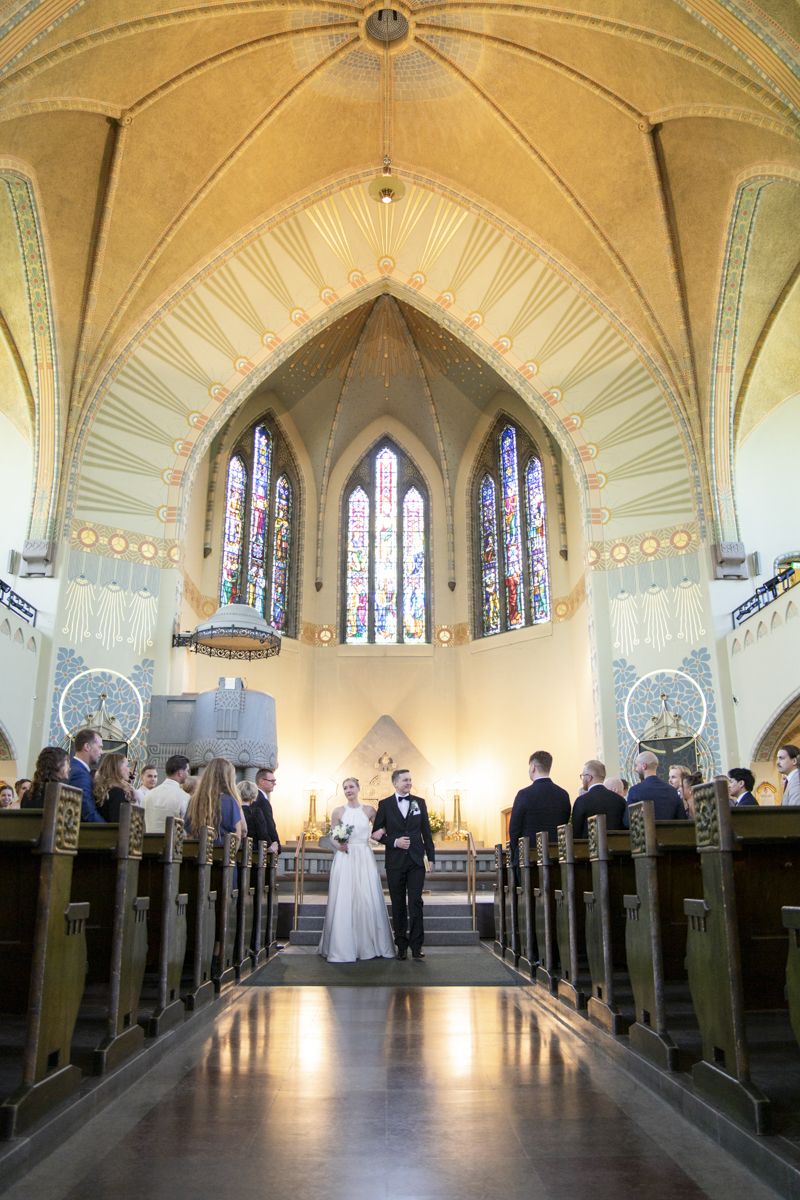 Bride and groom walking down the aisle inside a church with tall stained-glass windows and an ornate patterned ceiling while guests watch from wooden pews.
