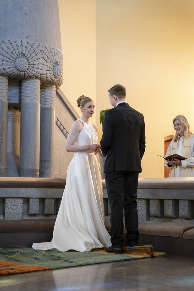 Bride in a white gown and groom in a black suit standing and holding hands in a church during wedding ceremony, with an officiant reading from a book behind an altar.
