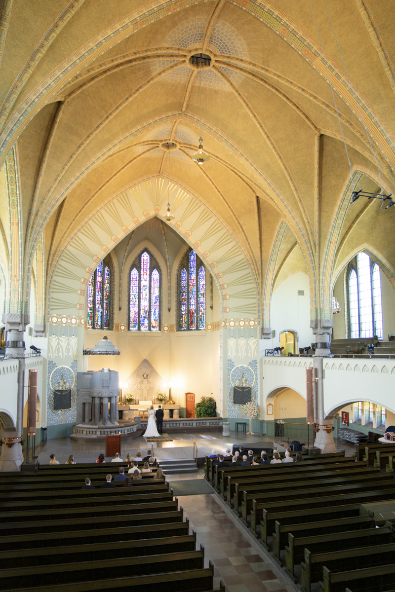 Interior of a church during a wedding ceremony with a bride and groom standing at the altar beneath a large vaulted ceiling and stained glass windows, with guests seated in wooden pews.