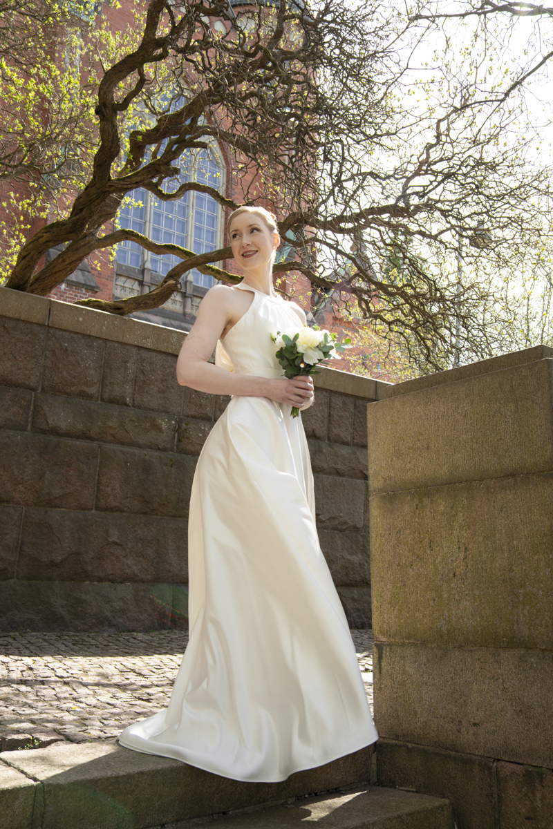 Bride in a white sleeveless wedding dress holding a bouquet of white flowers and standing on stone steps outdoors with a brick building and tree branches in the background.