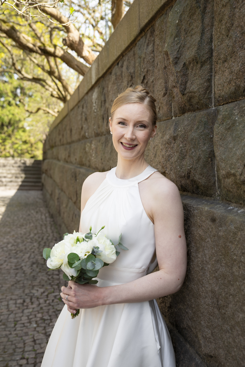 Smiling bride in a sleeveless white wedding dress holding a bouquet of white flowers and eucalyptus, standing against a stone wall outdoors with green trees in the background.
