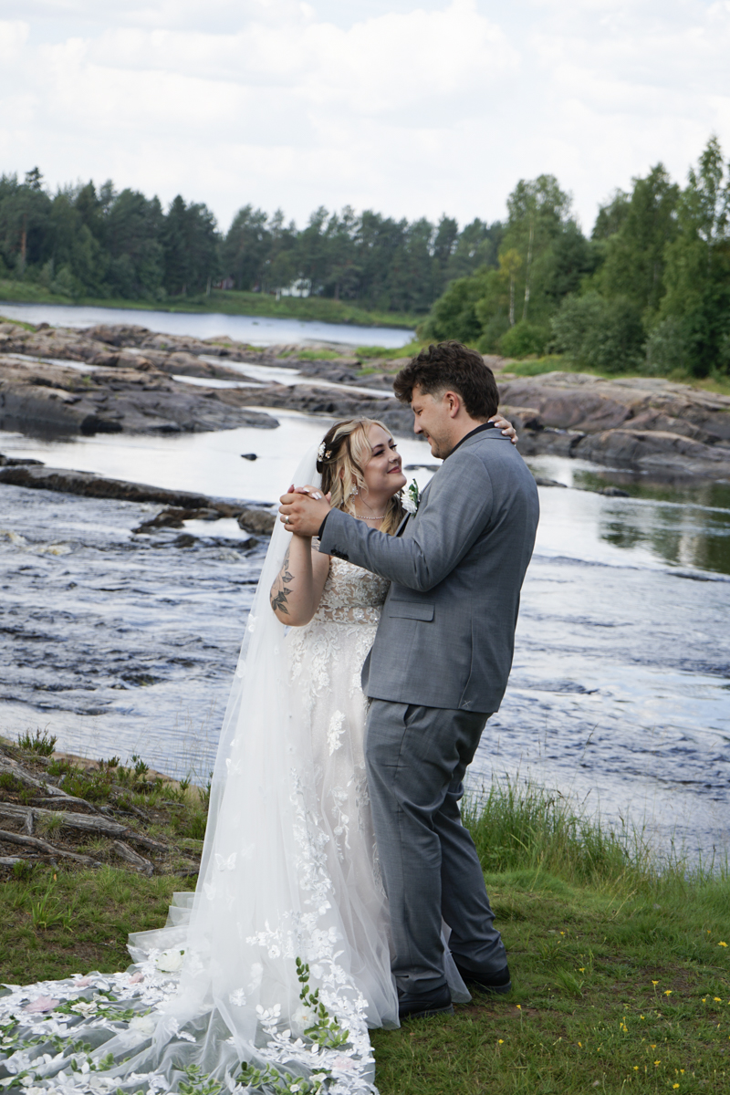 A bride and groom embracing and dancing closely by a rocky riverside with trees and greenery in the background on a cloudy day.
