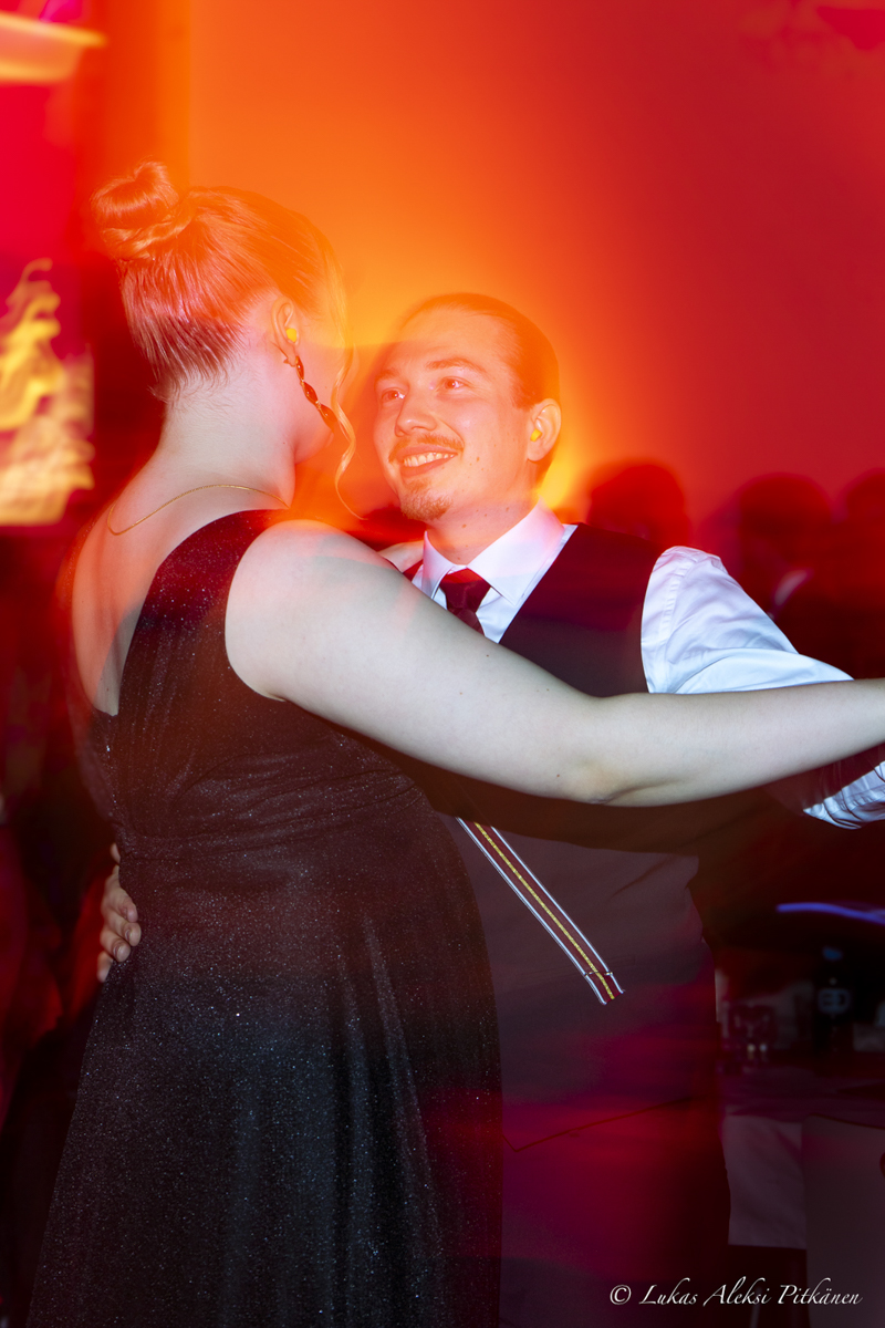 A couple dancing closely at a formal event with warm red and orange lighting around them; the man is smiling, wearing a white shirt, tie, and vest, and the woman has her hair in a bun and wears a black dress.