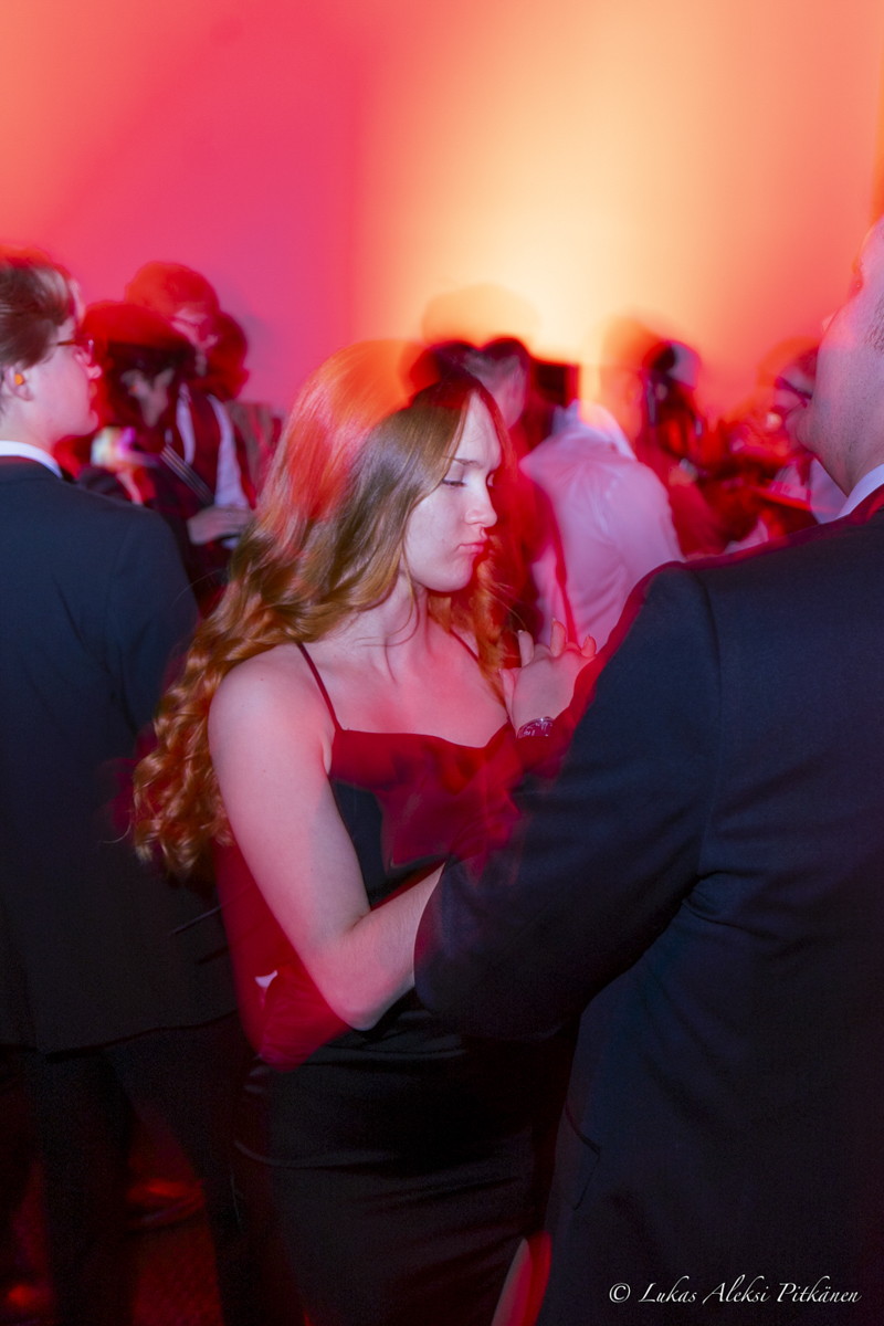 A young woman with long wavy hair in a sleeveless black dress dances closely with a man in a dark suit amid a dimly lit crowded party with red lighting effects.