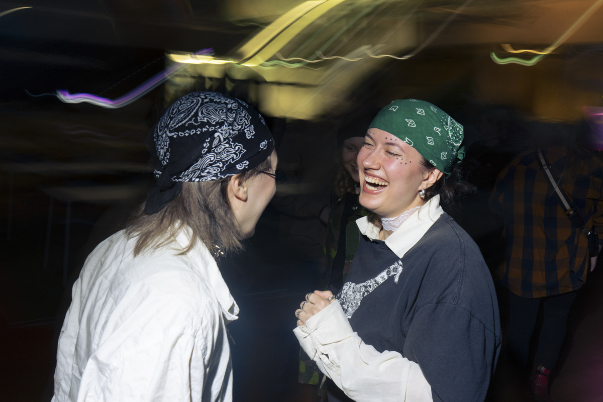 Two young people facing each other and laughing, both wearing bandanas—one black with a white pattern, the other green with a white pattern. One person wears glasses and a white shirt, the other wears a dark shirt over a white collared shirt and has small face decorations. The background is dimly lit with blurred lights and faint figures.