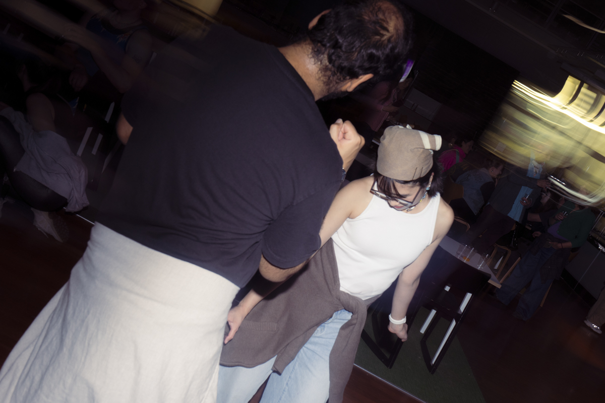 A man and woman dancing indoors at a social event; the man wears a black shirt and light-colored pants, the woman wears a white sleeveless top, light blue jeans, a brown sweater tied around her waist, and a brown hat.